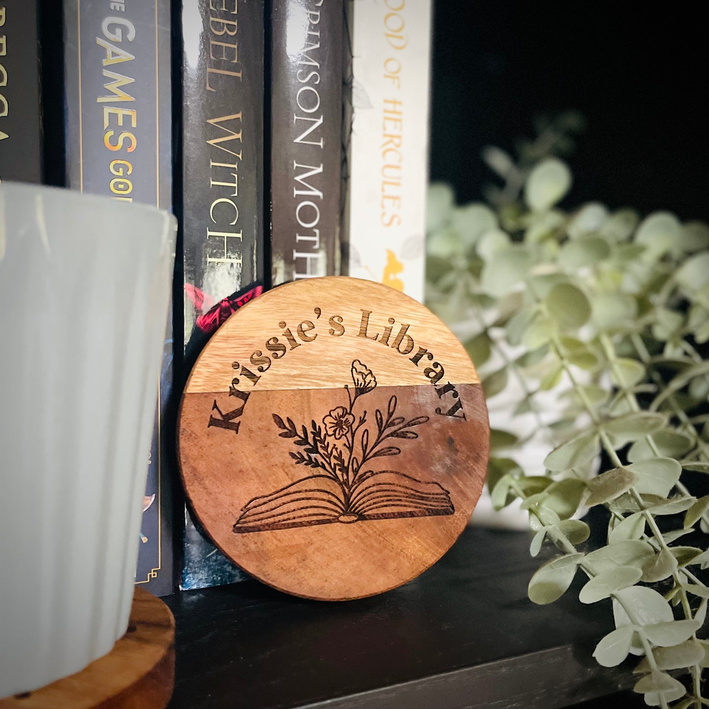 Wooden round sign with 'Krissie's Library' engraving on a bookshelf with books and plants.
