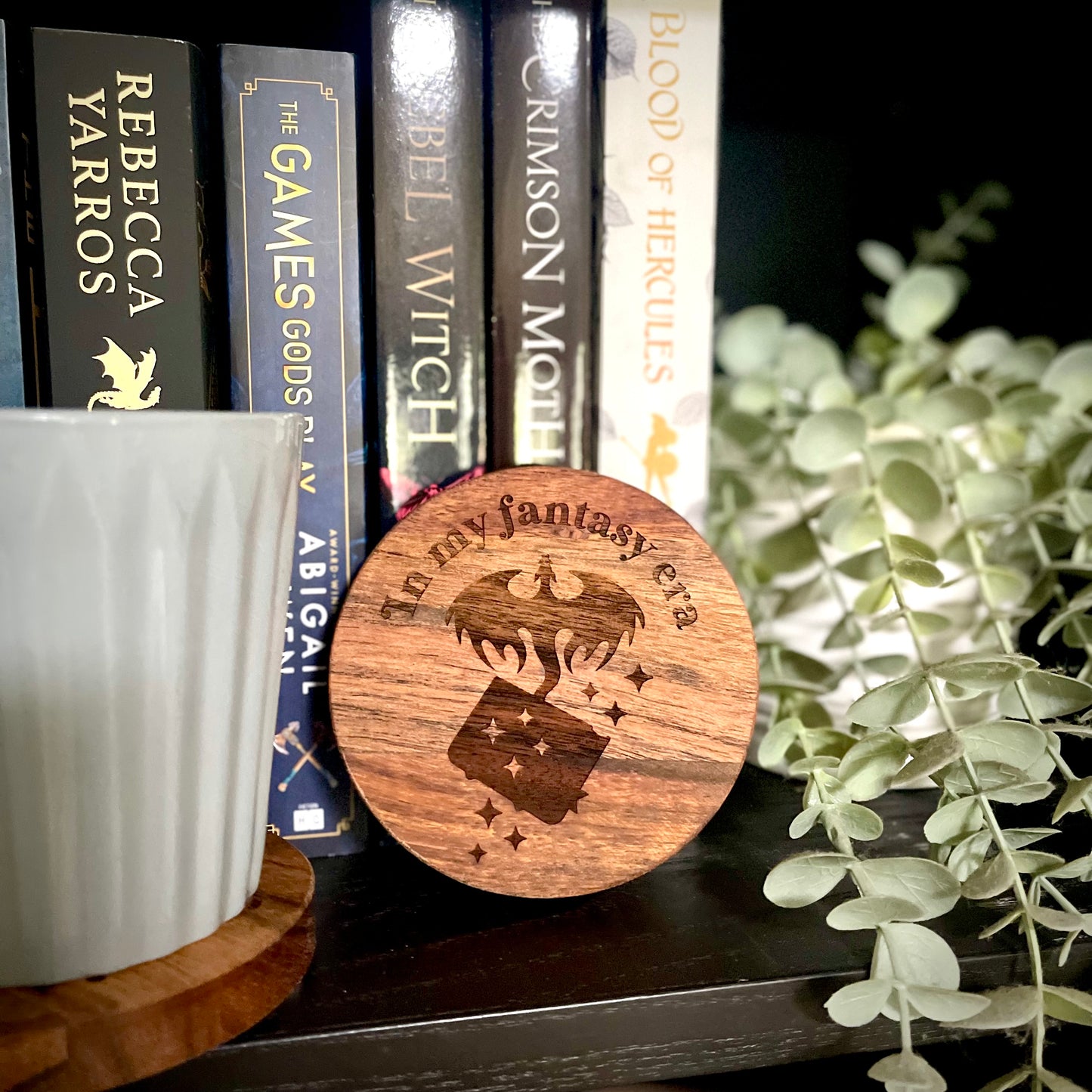 Wooden coaster with 'In my fantasy era' design on a bookshelf with books and a candle.