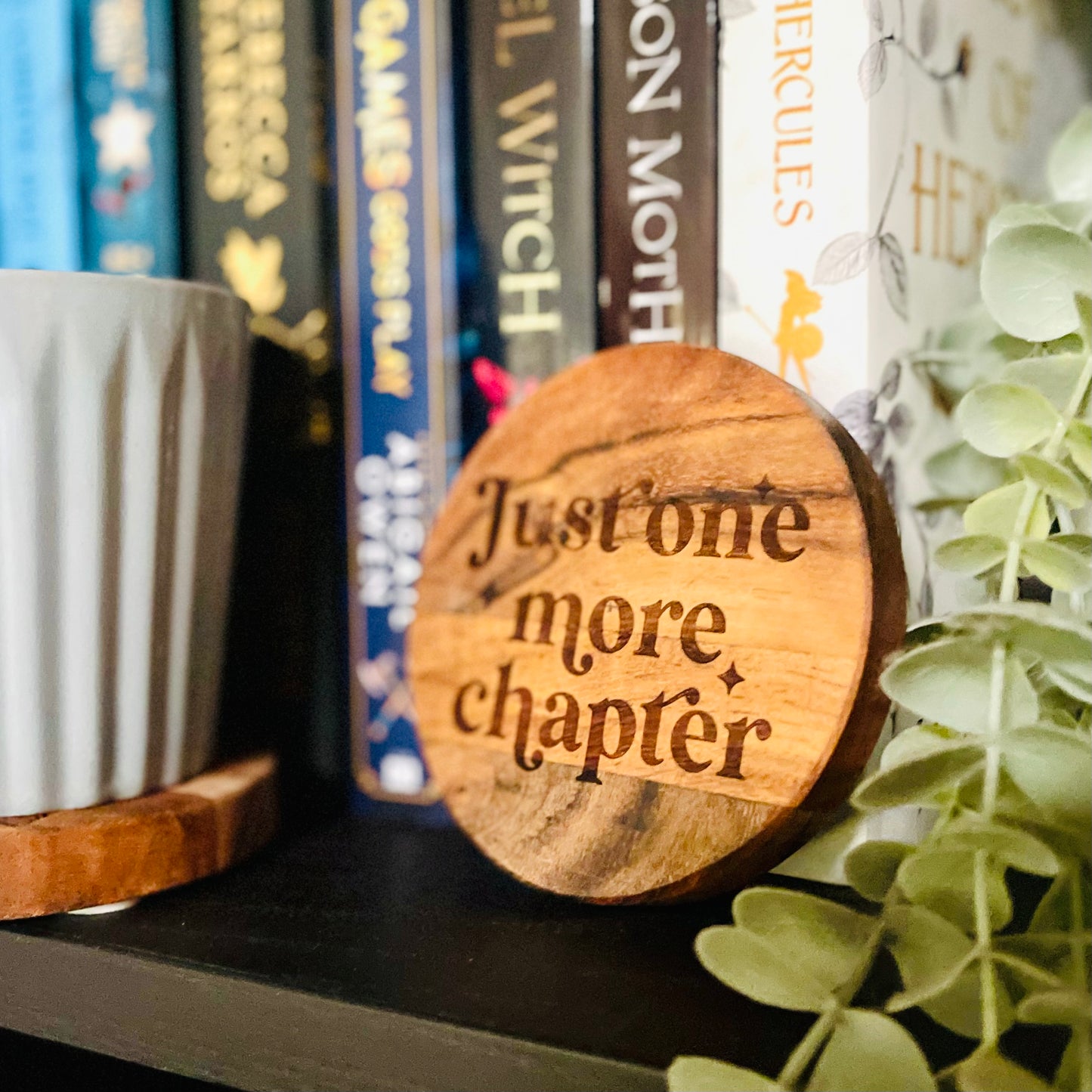Wooden coaster with 'Just one more chapter' text on a bookshelf with books and a plant.