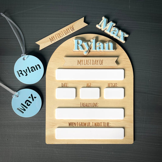 Wooden plaque with 'My First Day of School' and 'My Last Day of School' text, accompanied by two blue tags with names on a dark textured background.