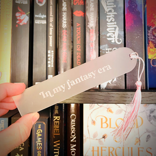Hand holding a 'In my fantasy era' bookmark in front of bookshelves with various books.