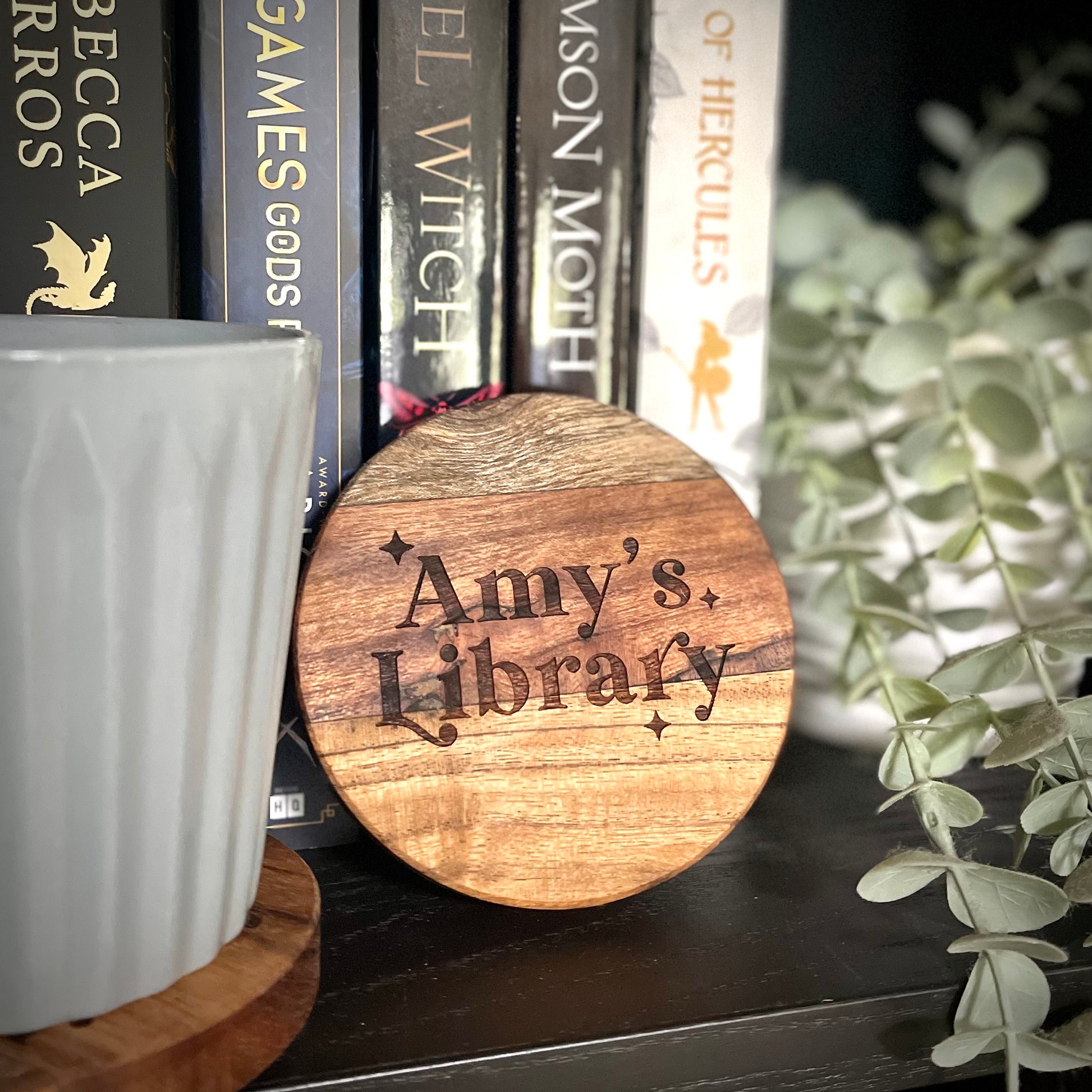 Wooden round sign with 'Amy's Library' engraved, placed on a shelf with books and a plant.