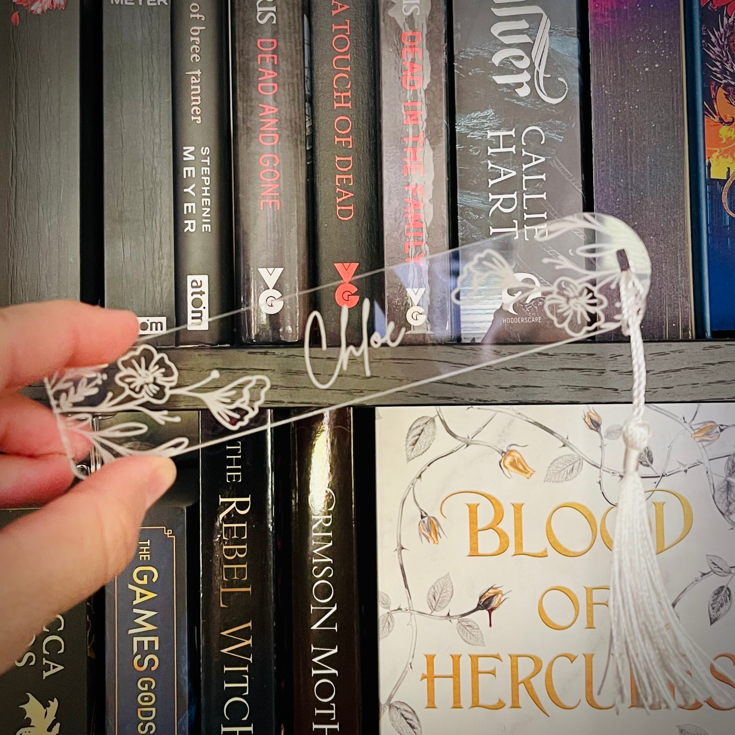 Hand holding a decorative bookmark in front of a bookshelf with books.