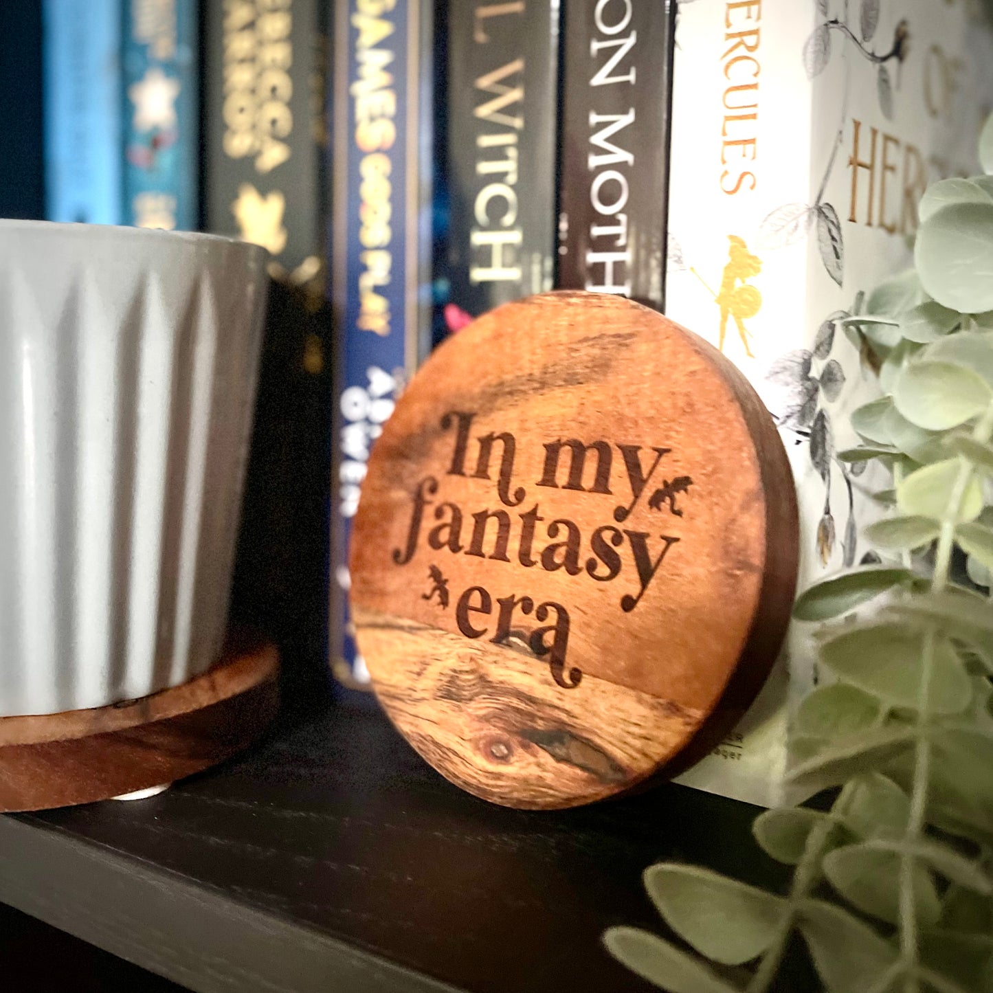 Wooden coaster with 'In my fantasy era' text on a bookshelf with books and plants.