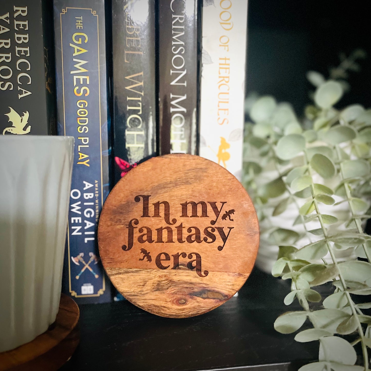 Wooden disc with 'In my fantasy era' text on a bookshelf with books and a plant.