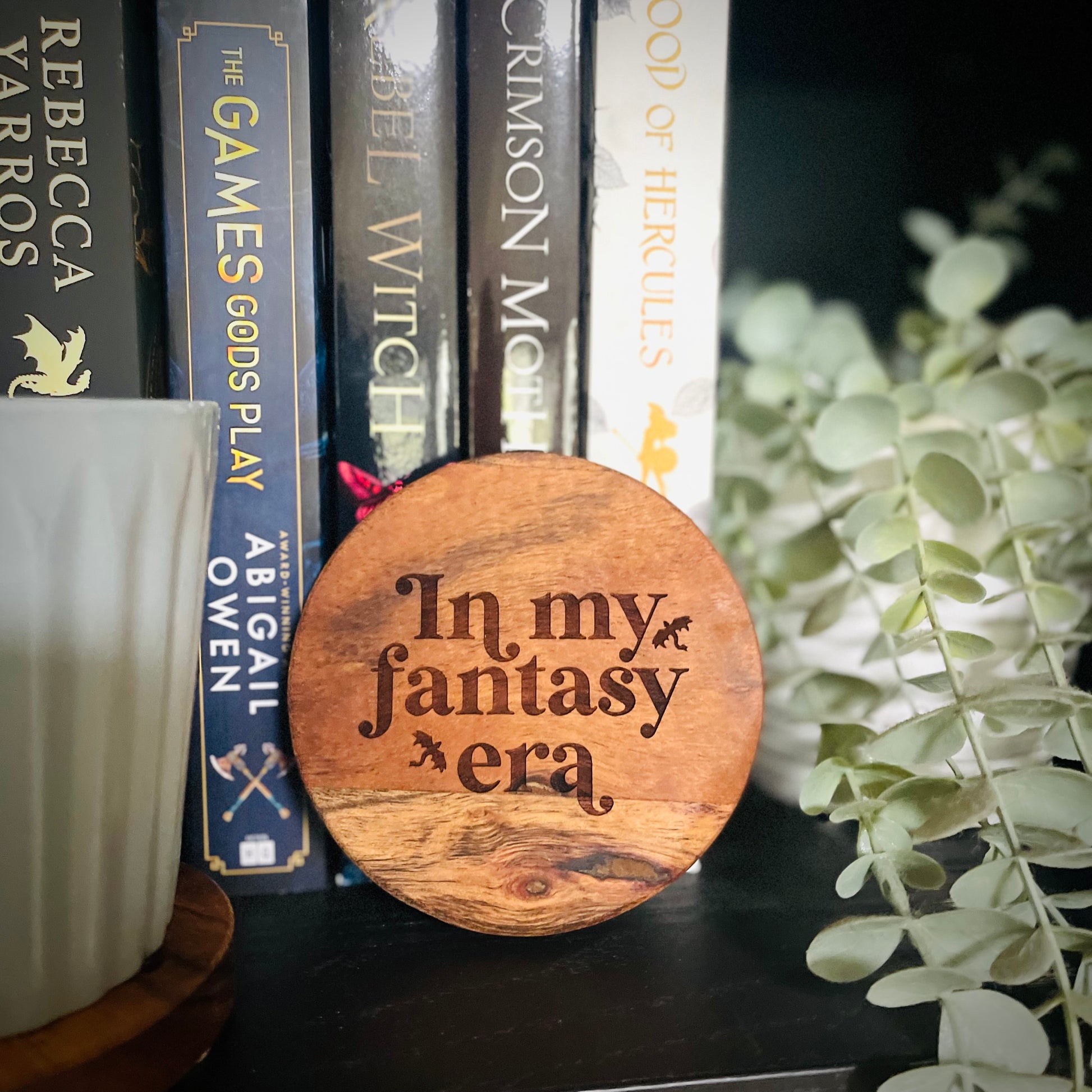 Wooden disc with 'In my fantasy era' text on a bookshelf with books and a plant.