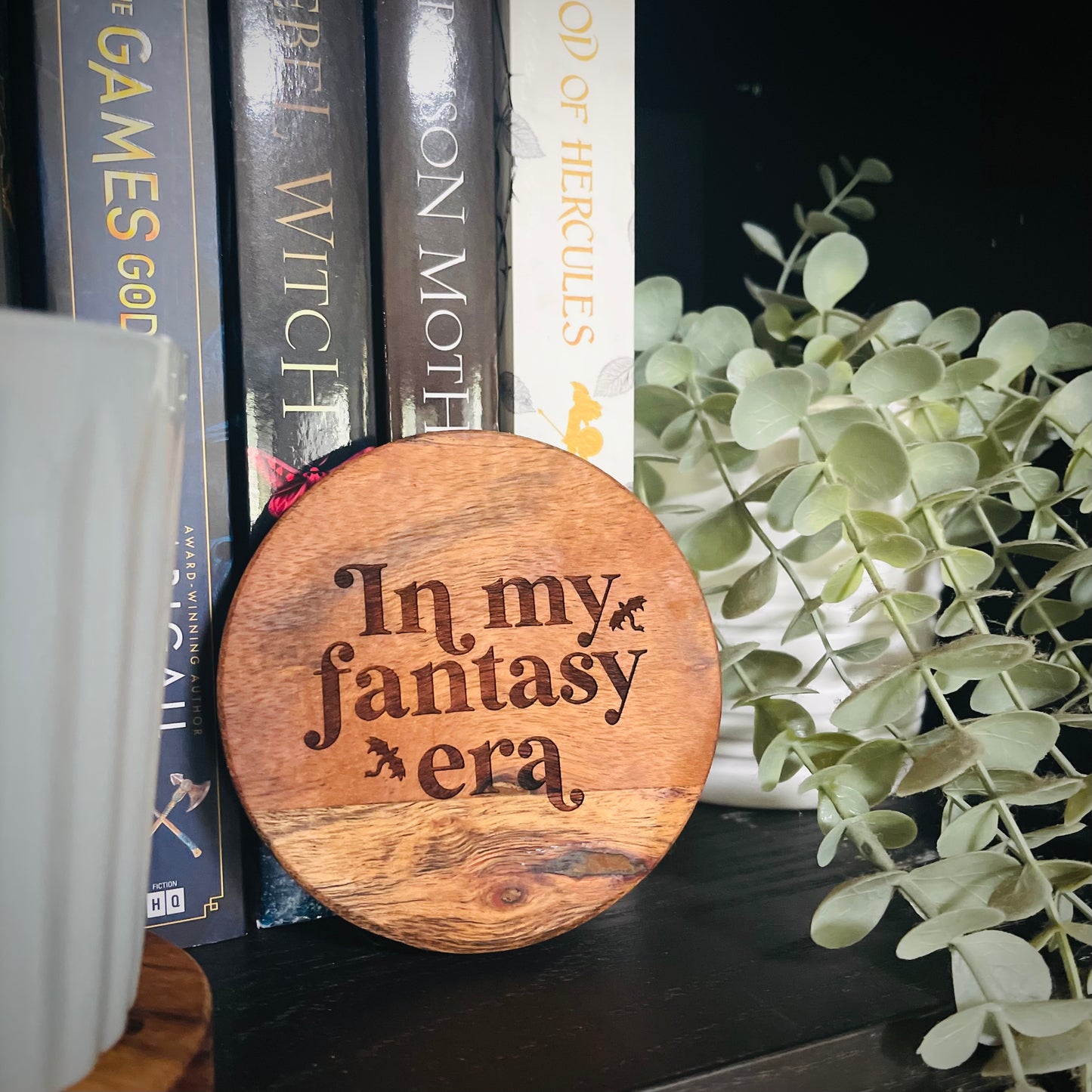 Wooden coaster with 'In my fantasy era' text on a bookshelf with books and plants.