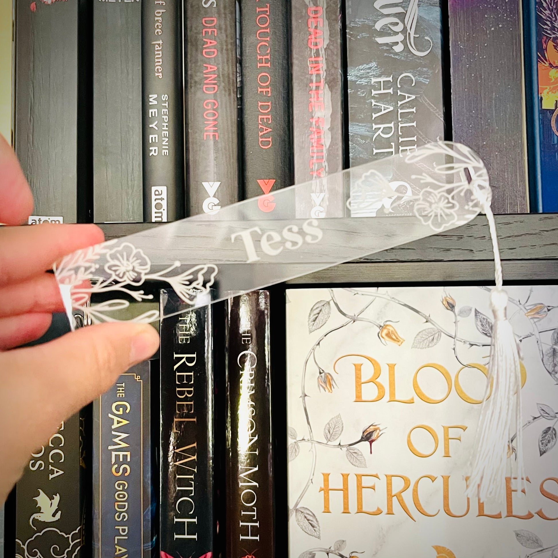 Hand holding a clear ribbon with 'Tess' on it in front of a bookshelf with books like 'The Games' and 'Blood of Hercules'.